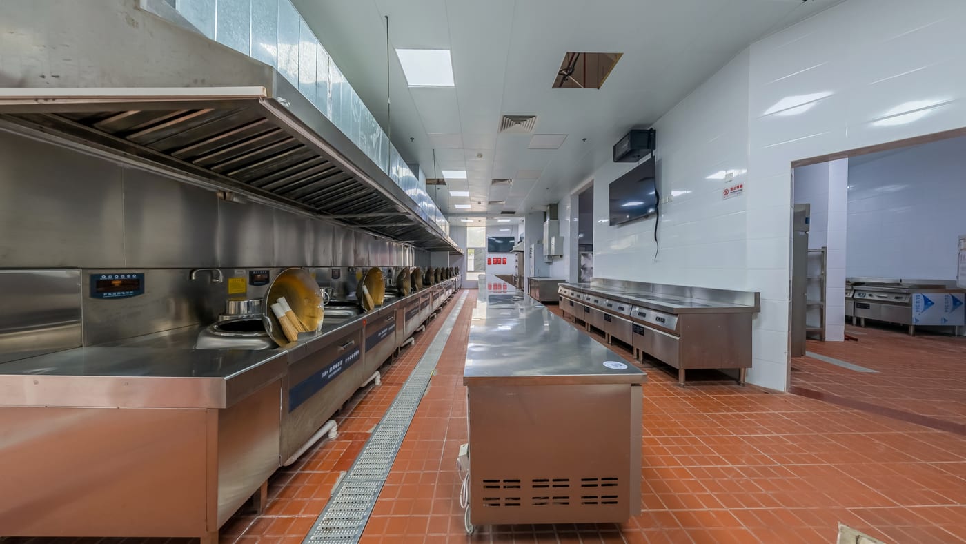 A long empty commercial kitchen, stainless steel counters and equipment down both sides, floor clear.
